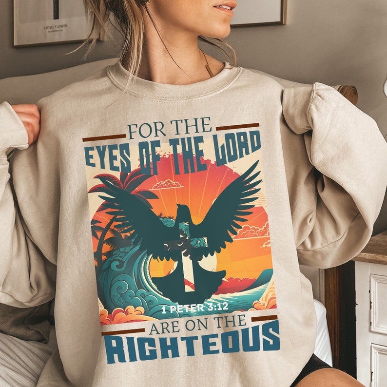 Puede incluir: Sudadera beige con un gr&aacute;fico de estilo vintage que presenta una paloma con una cruz, palmeras y una puesta de sol. El texto dice "Porque los ojos del Se&ntilde;or est&aacute;n sobre los justos" y "1 Pedro 3:12".
