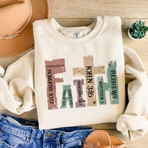 May include: A cream-coloured sweatshirt featuring the word "Faith" in a cross design. Each letter of "Faith" is a different colour and contains Bible verses: Ephesians 2:8, Romans 10:17, Psalm 37:5, James 2:17, Proverbs 3:5-6, John 3:16, Matthew 2, and Hebrews.