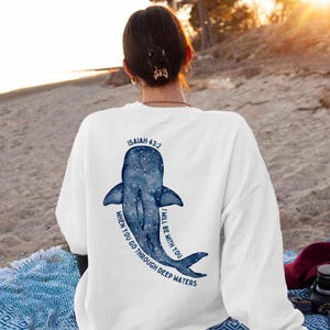 May include: White sweatshirt with a blue whale illustration and the text "ISAIAH 43:2" and the quote "WHEN YOU GO THROUGH DEEP WATERS, I WILL BE WITH YOU." The whale design is in a watercolor style.