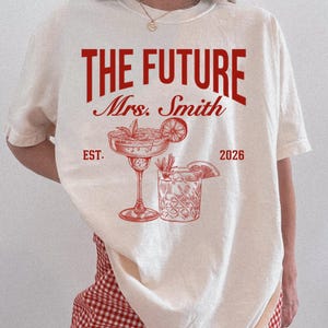 May include: Cream-colored t-shirt with the text "THE FUTURE Mrs. Smith EST. 2026" in red. The design includes illustrations of two cocktails. The shirt is a casual wear item, likely for a wedding or bridal event.