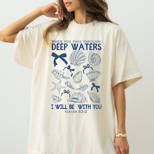 May include: A cream coloured t-shirt with a blue graphic design of seashells and bows. The text "WHEN YOU PASS THROUGH DEEP WATERS I WILL BE WITH YOU ISAIAH 43:2" is printed on the shirt.