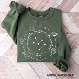 May include: Olive green sweatshirt with a white celestial design. The design includes planets, stars, and the text "THE HEAVENS DECLARE THE GLORY OF GOD: THE SKIES PROCLAIM THE WORK OF HIS HANDS. PSALM 19:1." The text "AVALIABLE IN HOODIE & TSHIRT" is at the bottom.