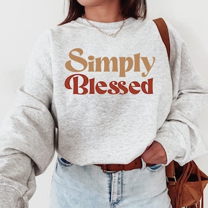 May include: A grey sweatshirt with the text "Simply Blessed" in a retro font. The text is in shades of brown and red.