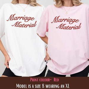 Puede incluir: Dos camisetas holgadas, una blanca y otra rosa, cada una con la frase "Marriage Material" en letras rojas. Las camisetas se combinan con pantalones cortos negros. El texto "Print Colour - Red" y "Model is a size 8 wearing an XL" est&aacute; en la parte inferior.