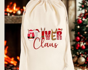 May include: A cream-colored drawstring gift bag with the words "Oliver Claus" in red script and festive lettering. The word "Oliver" is decorated with reindeer and Santa hat designs, perfect for Christmas.