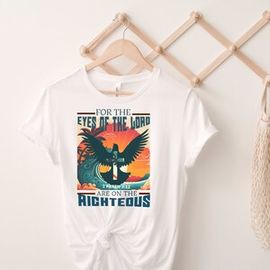 Puede incluir: Camiseta blanca con un dise&ntilde;o gr&aacute;fico que presenta una paloma con las alas extendidas volando sobre una cruz. El gr&aacute;fico est&aacute; situado sobre un fondo de puesta de sol con palmeras y olas. El texto "For the Eyes of the Lord Are on the Righteous" est&aacute; impreso debajo del gr&aacute;fico, junto con la referencia b&iacute;blica "1 Pedro 3:12".