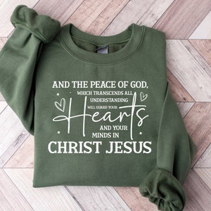 以下が含まれることがあります： 白い文字で「AND THE PEACE OF GOD, WHICH TRANSCENDS ALL UNDERSTANDING WILL GUARD YOUR Hearts AND YOUR MINDS IN CHRIST JESUS」と書かれた緑色のスウェットシャツ。