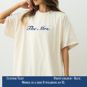 May include: Cream-coloured oversized t-shirt with the text "The Mrs." in navy blue. The shirt has short sleeves and a relaxed fit. The text is centered on the chest.