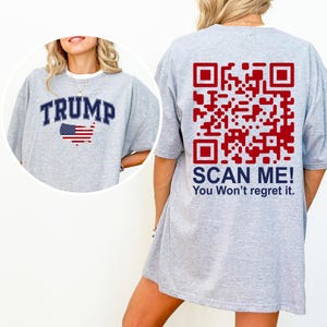 Może przedstawiać: Szara koszulka z napisem "TRUMP" w niebieskich literach i amerykańską flagą z przodu. Z tyłu koszulki znajduje się kod QR z tekstem "SCAN ME! You Won't regret it."