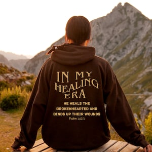 Op de afbeelding: Een bruine hoodie met de tekst "IN MY HEALING ERA" en het citaat "HE HEALS THE BROKENHEARTED AND BINDS UP THEIR WOUNDS" met de bijbelverwijzing "Psalm 147:3".