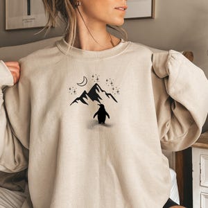 Op de afbeelding: Beige sweatshirt met een zwarte afbeelding van een pingu&iuml;n voor een bergketen, een maan en sterren. Het ontwerp is gecentreerd op de voorkant van de top met lange mouwen en ronde hals.