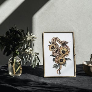 Puede incluir: Una impresi&oacute;n enmarcada de una pintura de acuarela que presenta una serpiente marr&oacute;n y blanca envuelta alrededor de flores amarillas. La serpiente est&aacute; enroscada alrededor de las flores, con la cabeza mirando al espectador. Las flores est&aacute;n en flor, con p&eacute;talos amarillos y hojas verdes. La impresi&oacute;n est&aacute; enmarcada en un marco dorado.