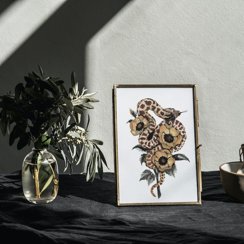 Puede incluir: Una impresi&oacute;n enmarcada de una pintura de acuarela que presenta una serpiente marr&oacute;n y blanca envuelta alrededor de flores amarillas. La serpiente est&aacute; enroscada alrededor de las flores, con la cabeza mirando al espectador. Las flores est&aacute;n en flor, con p&eacute;talos amarillos y hojas verdes. La impresi&oacute;n est&aacute; enmarcada en un marco dorado.