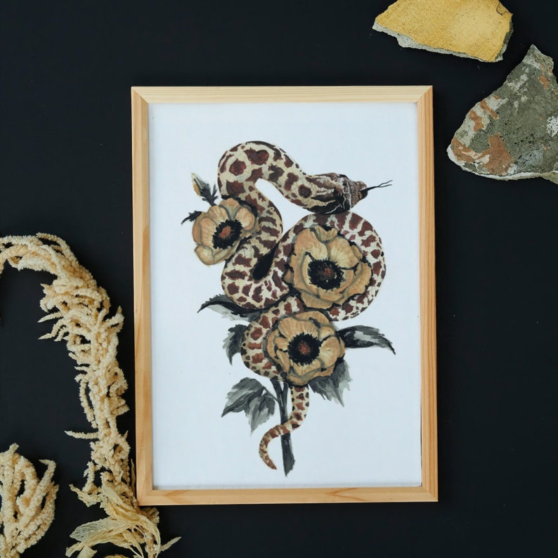 Puede incluir: Una impresi&oacute;n enmarcada de una pintura de acuarela que presenta una serpiente marr&oacute;n y blanca enroscada alrededor de tres flores amarillas con centros marrones. Las flores tienen hojas y tallos verdes.
