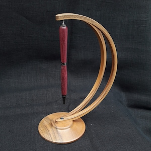 May include: A handcrafted pen display stand made of wood, holding a burgundy pen. The stand has a curved, arching design with a circular base. The pen is suspended in mid-air, showcasing its design.
