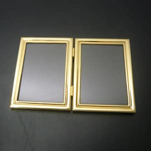 Puede incluir: Un portafotos doble articulado, con acabado dorado. Cada marco rectangular tiene un interior gris oscuro. Los marcos están conectados por una bisagra dorada, lo que permite plegarlos. Un artículo decorativo.