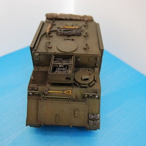 May include: An olive drab military armoured personnel carrier scale model viewed from above. The vehicle features a flat roof with a circular hatch, a rear cargo area with a shovel, and various moulded details including storage compartments and tools.