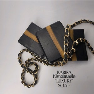 May include: Three black and gold soap bars with a gold chain on top. The soap bars have a black base with a gold stripe running through the center. The chain is made of gold links with black accents. The text "KARINA_handmade" and "LUXURY SOAP" is visible below the soap bars.