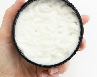 May include: A black container filled with a thick, white cream. The cream has a smooth, textured appearance. The container is held in a person's hand against a white background. The cream appears to be a skincare product.