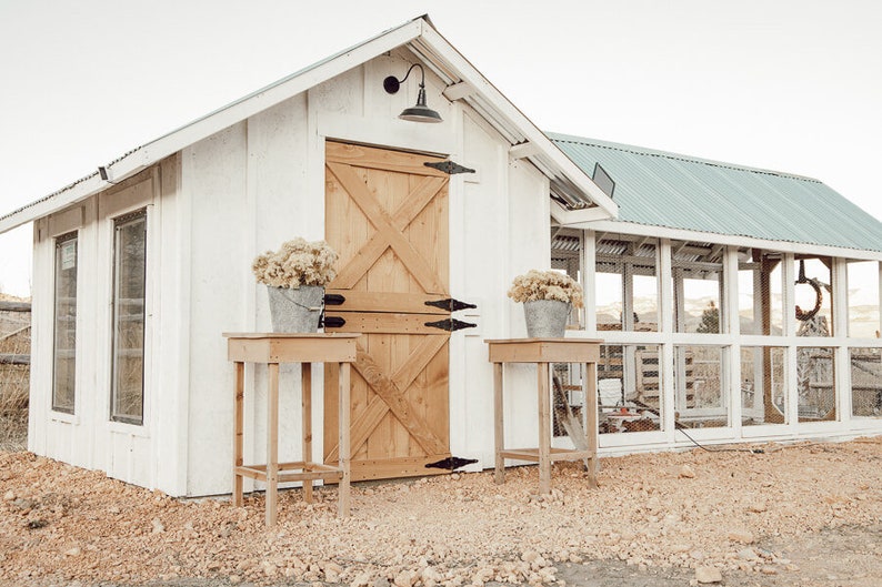 May include: A white wooden shed with a light brown wooden door and a light brown wooden table. Two galvanized metal buckets with dried flowers sit on the tables. The shed has a black light fixture and a green metal roof.