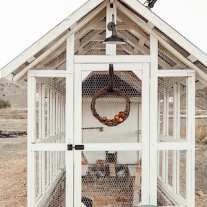 May include: A white wooden chicken coop with wire mesh panels and a gabled roof. A decorative wreath hangs inside the coop, and a metal container sits in front. A black light fixture is mounted above the coop.