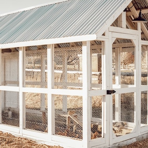 May include: A white wooden chicken coop with a corrugated metal roof. The coop has wire mesh panels and a black light fixture. Chickens are visible inside the coop. The coop is outdoors, with a fence and trees in the background.