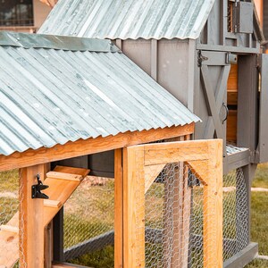 May include: A gray and natural wood chicken coop with a corrugated metal roof. The coop has a hinged door with wire mesh and a small window. The coop is constructed with wooden framing and is set on a grassy lawn.