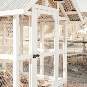 May include: A white wooden chicken coop with a corrugated metal roof. The coop features wire mesh walls and a small door with a black latch. A black light fixture is mounted on the side. The coop is set on a gravel surface.