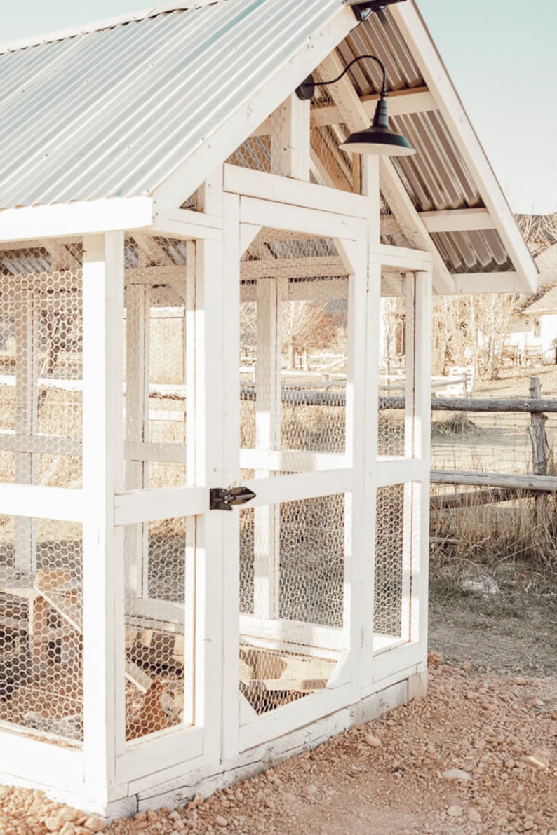 May include: A white wooden chicken coop with a corrugated metal roof. The coop features wire mesh walls and a small door with a black latch. A black light fixture is mounted on the side. The coop is set on a gravel surface.