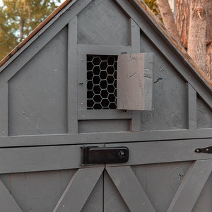 May include: A gray wooden structure with a gabled roof, featuring a small window with wire mesh and a hinged door. The structure has a black latch and handles, and is set against a backdrop of trees.