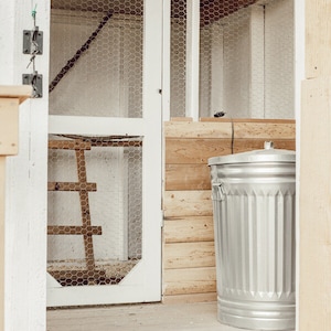 May include: A silver metal trash can with vertical ridges stands next to a wooden structure. The structure has a white door with wire mesh and a wooden ladder inside. The scene is set on a wooden floor.
