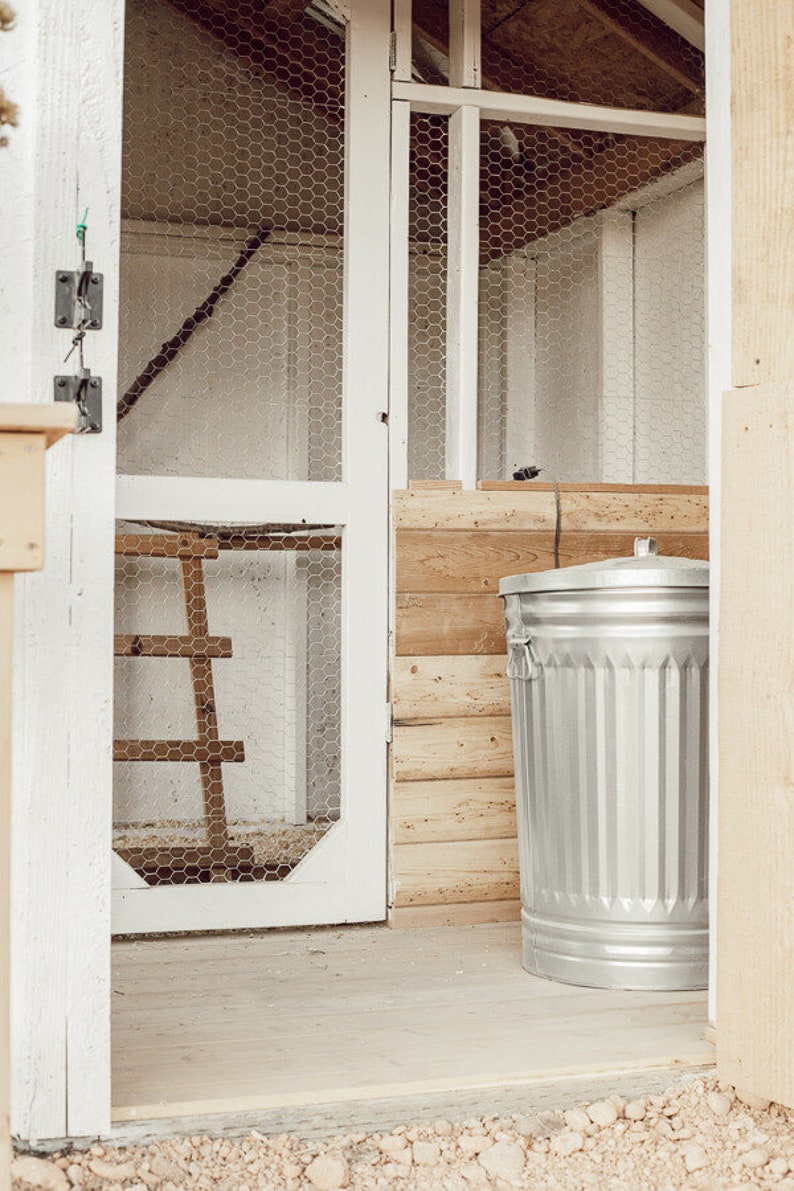 May include: A silver metal trash can with vertical ridges stands next to a wooden structure. The structure has a white door with wire mesh and a wooden ladder inside. The scene is set on a wooden floor.