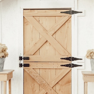 May include: A rustic wooden door with an X-shaped design, black hinges, and a white frame. Two small wooden tables flank the door, each holding a galvanized bucket filled with dried flowers. The scene evokes a farmhouse aesthetic.