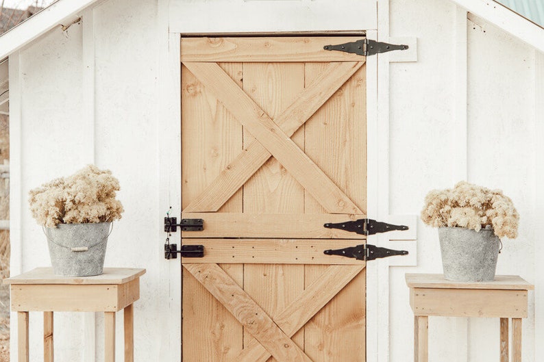 May include: A rustic wooden door with an X-shaped design, black hinges, and a white frame. Two small wooden tables flank the door, each holding a galvanized bucket filled with dried flowers. The scene evokes a farmhouse aesthetic.