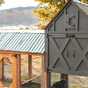 May include: A wooden chicken coop with a gray exterior and a corrugated metal roof. The coop features a door with black handles and a wire mesh enclosure. Chickens are visible inside the coop, with a scenic background of trees and a mountain.