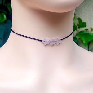 May include: A delicate choker necklace featuring three pale purple star-shaped beads strung on a thin, dark blue cord. The necklace is displayed on a mannequin, showcasing its design and style. The background is blurred, with green foliage.