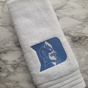 May include: White hand towel with a blue and white embroidered logo. The logo features a blue shield with a white illustration of a devil. The towel has a striped border at the bottom.