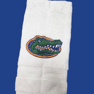 May include: White hand towel with an embroidered green alligator logo inside an orange and blue oval. The towel has a textured design near the bottom and is set against a blue background.