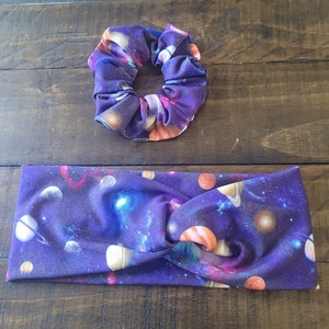 May include: A matching set of hair accessories featuring a galaxy print. The set includes a scrunchie and a twisted headband. The fabric is a deep purple with colorful planets, stars, and nebulae.