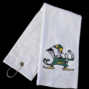 May include: White golf towel with a green and gold embroidered leprechaun graphic. The towel has a metal loop for hanging. The leprechaun is wearing a green outfit and hat.