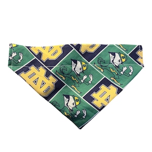 May include: Green, navy, and gold dog bandana featuring a repeating pattern of the Notre Dame Fighting Irish leprechaun mascot and the letters "ND". The bandana is triangular in shape and made of fabric.