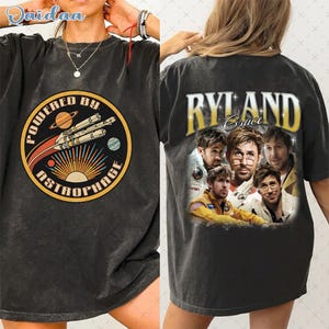 Puede incluir: Camiseta gris oscuro con un diseño gráfico retro en la parte delantera con el texto "POWERED BY ASTROPHAGE" y un diseño de cohete. La parte trasera de la camiseta muestra el texto "RYLAND Grace" con un collage de imágenes.