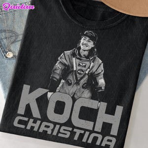 Puede incluir: Camiseta negra con un retrato de Christina Koch con un traje espacial de la NASA. El nombre "KOCH CHRISTINA" está impreso en letras grises grandes y desgastadas debajo de la imagen. La camiseta tiene cuello redondo y mangas cortas.