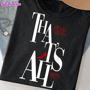Op de afbeelding: Zwart T-shirt met de tekst "THAT'S ALL" in witte letters. De woorden "THE DEVIL WEARS PRADA" zijn rood, met een rode afbeelding van een schoen met hoge hakken. De tekst "-MIRANDA PRIESTLY" staat onderaan.