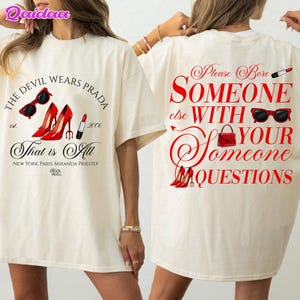 May include: Off-white t-shirt featuring the text "THE DEVIL WEARS PRADA" and "That is All" in black. The design includes red high heels, sunglasses, and lipstick. The back of the shirt says "Please Bore SOMEONE else WITH YOUR QUESTIONS".