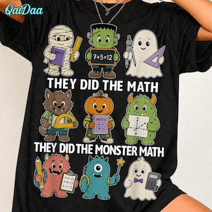 May include: A black t-shirt featuring a graphic print of nine cartoon monsters holding math tools and books. The text reads "THEY DID THE MATH" and "THEY DID THE MONSTER MATH". The characters include a mummy, Frankenstein, ghosts, a werewolf, a pumpkin-headed figure, and other colorful creatures.