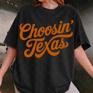 May include: A black short-sleeved t-shirt featuring the text Choosin' Texas printed in a large, bold, orange cursive font across the chest. The garment has a crew neckline and a relaxed fit, showcasing a simple design with high-contrast lettering.