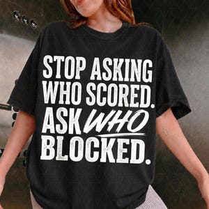 May include: A black short-sleeved t-shirt featuring white distressed-style text that reads: STOP ASKING WHO SCORED. ASK WHO BLOCKED. The shirt has a crew neckline and a relaxed fit, with the bold lettering centred prominently on the front chest area.