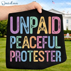 May include: Black protest sign with the words "UNPAID PEACEFUL PROTESTER" in colorful, distressed lettering. The sign is held up in front of the White House, with a fountain and American flag visible in the background.