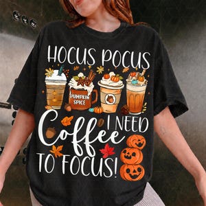 May include: A black t-shirt featuring a graphic of four coffee drinks, autumn leaves, and pumpkins. The text reads HOCUS POCUS I NEED COFFEE TO FOCUS! The design includes illustrations of mugs and cups with whipped cream, cinnamon sticks, and acorns.